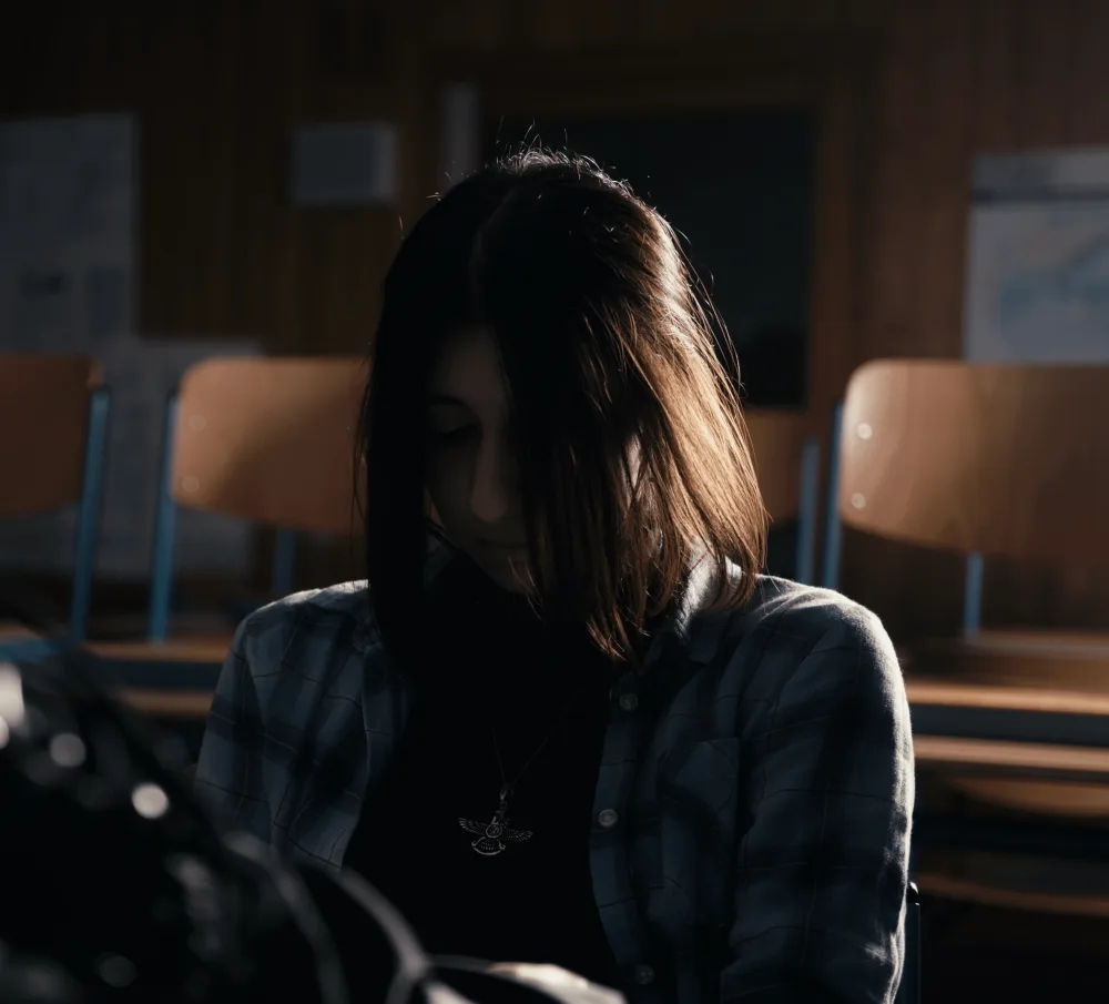 Filmstill einer einsamen Person, die allein an einem Schultisch im Klassenzimmer sitzt, die Szene wirkt ruhig und melancholisch.