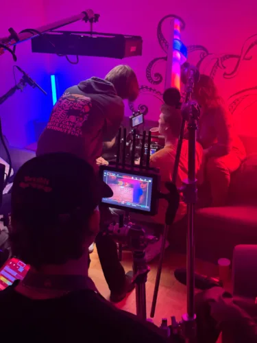 Indie-Filmset mit intensiver, farbiger Beleuchtung. Mehrere Teammitglieder arbeiten konzentriert an einer Kamera, während Schauspieler auf einem Sofa sitzen. Mikrofone und Filmtechnik sind gut sichtbar.