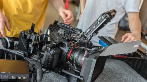 Verschiedene Kamera- und Filmequipment liegen auf einem Tisch bereit zur Vorbereitung eines Drehs. Im Hintergrund sind Crewmitglieder bei der Arbeit zu sehen.