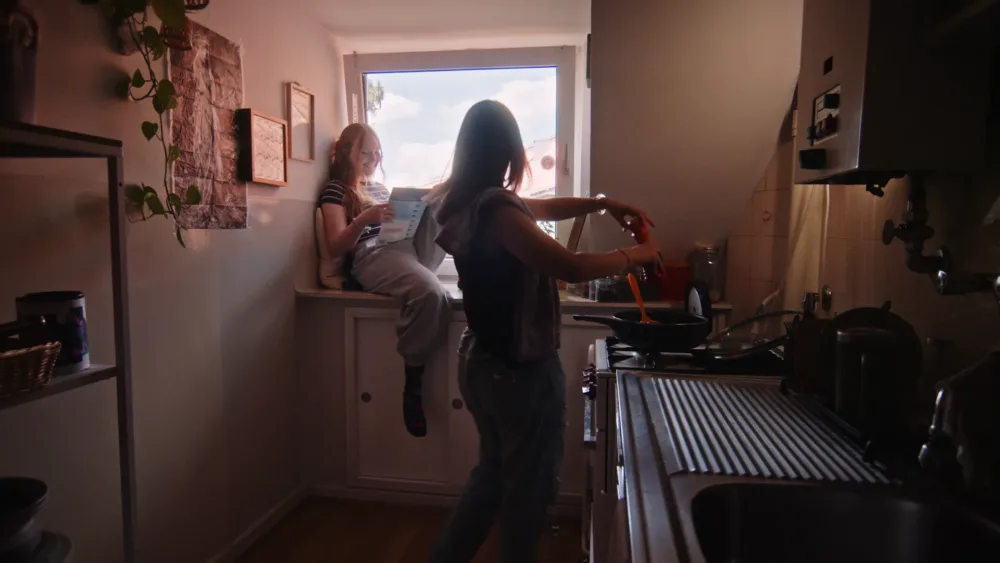 Ein stimmungsvoller Film-Still zeigt zwei Frauen in einer Küche: Eine sitzt lesend auf der Fensterbank, die andere kocht am Herd. Das Bild fängt einen intimen, authentischen Moment des Alltags ein.