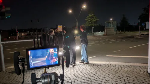 Nachtdreh an einer Straßenecke. Drei Personen stehen unter Straßenlaternen, während im Vordergrund ein Kameramonitor die Szene zeigt. Die Atmosphäre ist von künstlichem Licht und Dunkelheit geprägt.