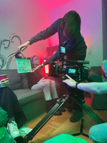 Dreharbeiten in einem Wohnzimmer mit bunter Beleuchtung. Ein Mitglied des Filmteams hält eine Filmklappe vor eine Schauspielerin, während eine Kamera auf einem Stativ die Szene aufnimmt.