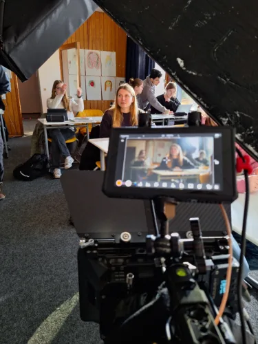 Blick hinter die Kulissen eines Filmdrehs in einem Klassenzimmer. Im Vordergrund ist ein Kameramonitor zu sehen, der eine Schülerin zeigt, die an ihrem Platz sitzt. Im Hintergrund arbeiten weitere Schüler und das Filmteam.
