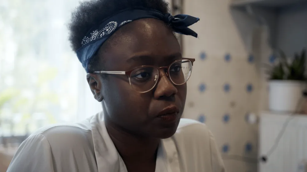 Filmstill: Eine Frau mit ausdrucksvollem Gesicht, Brille und blauem Kopftuch, in einer Innenaufnahme.